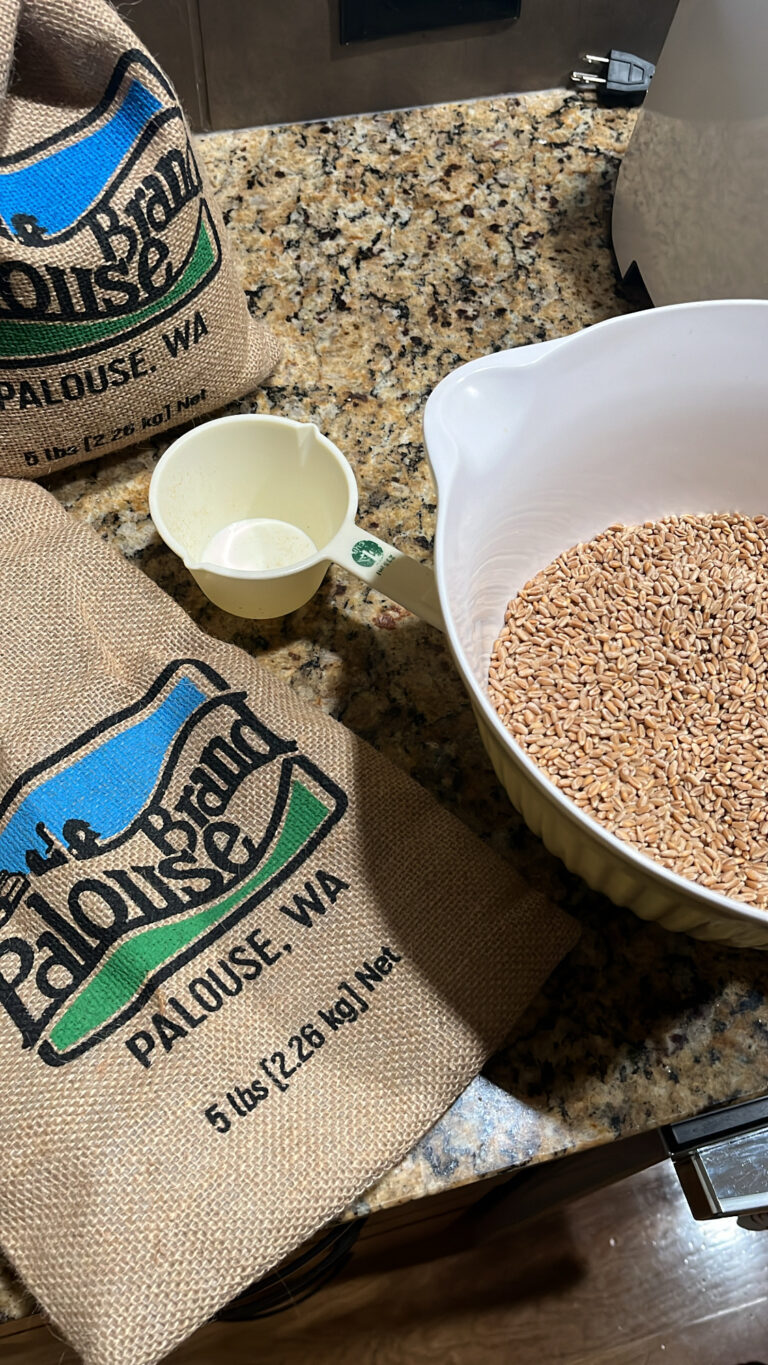 bags of palouse grain on a brown speckled granite counter top with a measuring cup, bowl with grain and partial mill in the picture